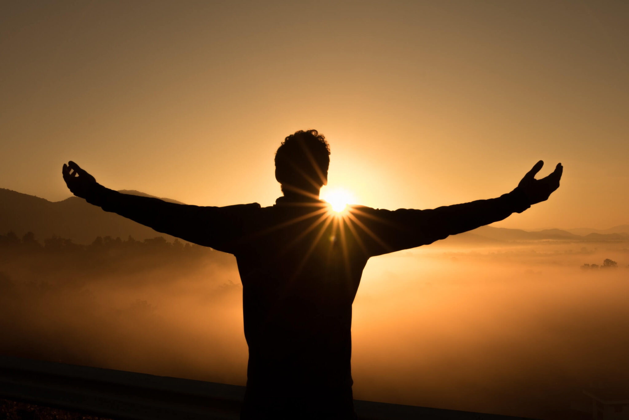 A man standing with his arms wide open facing a setting sun.
