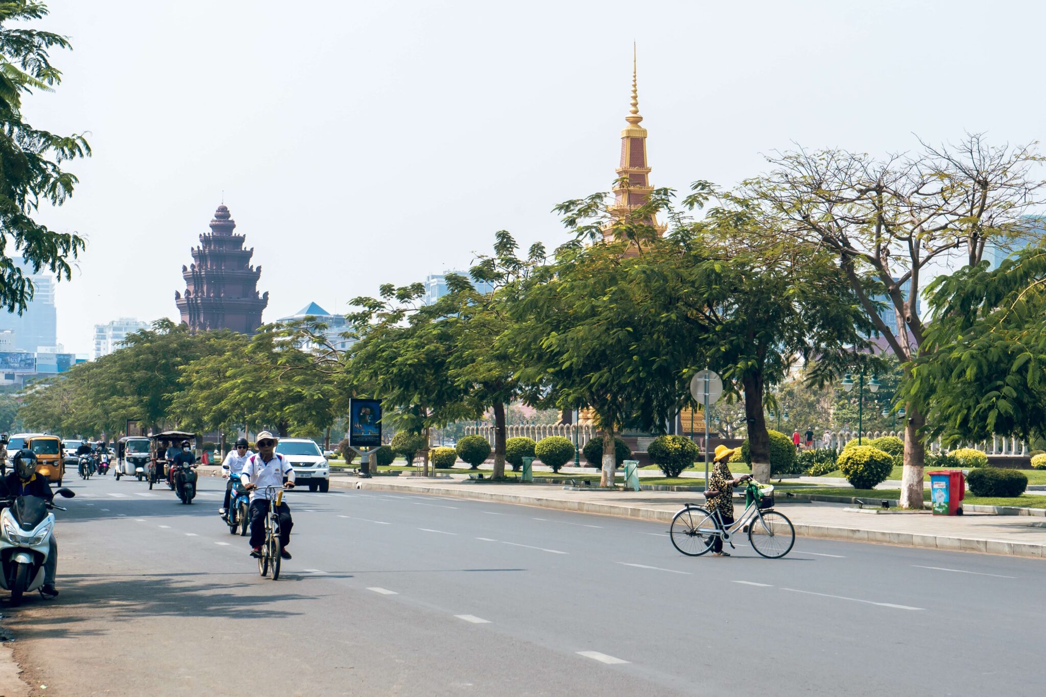 A city street in Cambodia - people are biking and a few cars are driving.