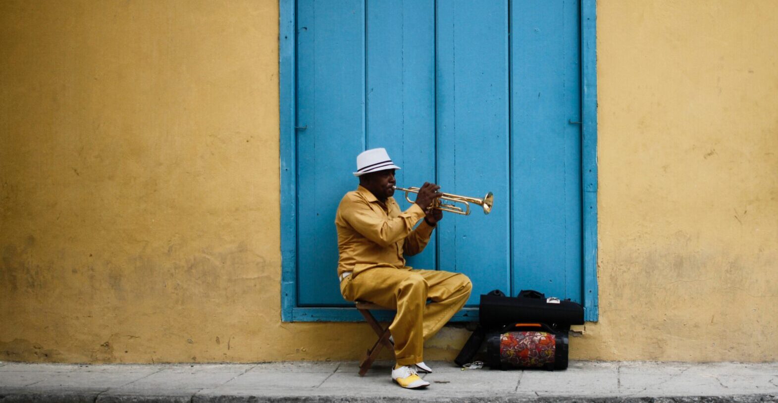 A man is sitting on a stool in front of a blue door playing a trumpet.