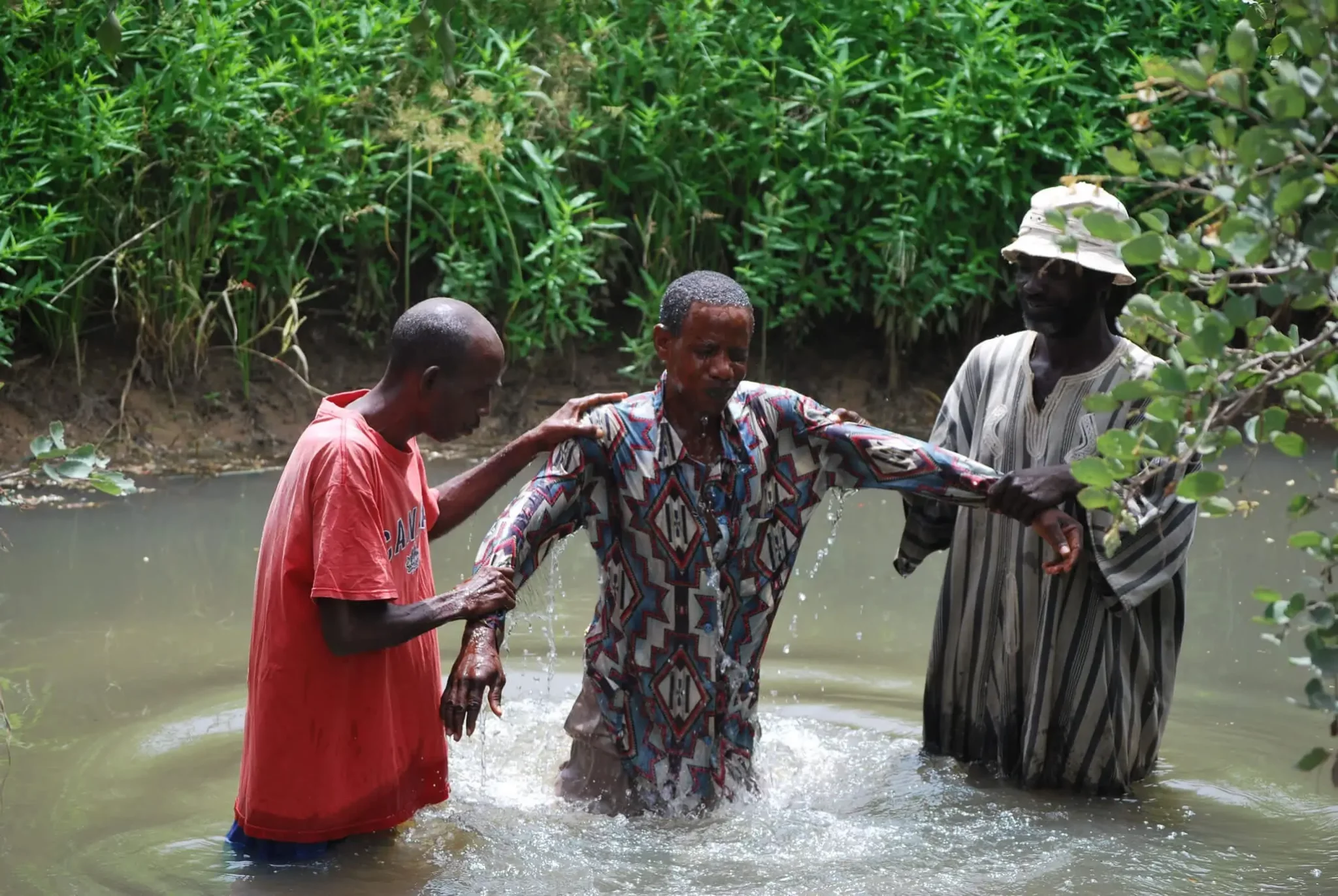 Two men baptizing a man in the jungle. The man in the middle was baptized and is emerging from the water with help from the other two men.