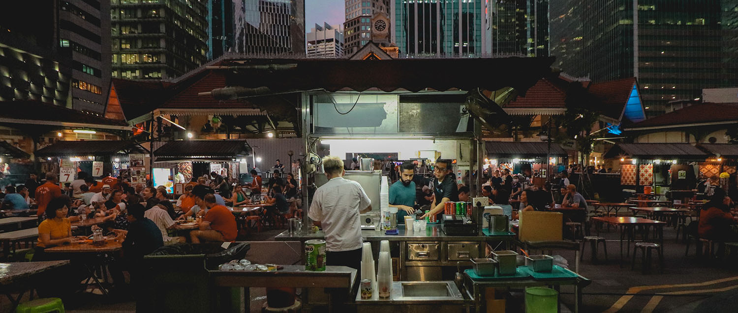 A picture of hawker stalls in southeast asia. People are sitting at small tables, eating, chatting, and surrounded by food vendors serving snacks from small carts.