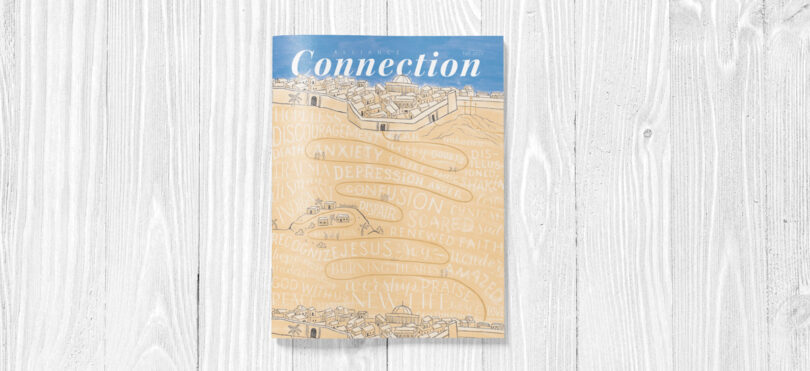Mockup of the Fall 2022 Alliance Connection magazine on a white wood background. A hand-lettered depiction of the Emmaus Road.